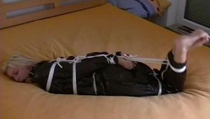 Blond-haired maid tied and gagged on bed wearing a shiny black PVC sauna suit (Video)