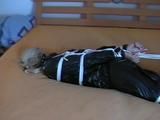 Blond-haired maid tied and gagged on bed wearing a shiny black PVC sauna suit (Video)