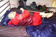 Pia tied and gagged on bed wearing a shiny red rain pants and a shiny red/purple down jacket (Pics)