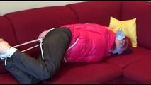 Mara tied and gagged on a sofa wearing a shiny pink down jacket and a black rain pants (Video)